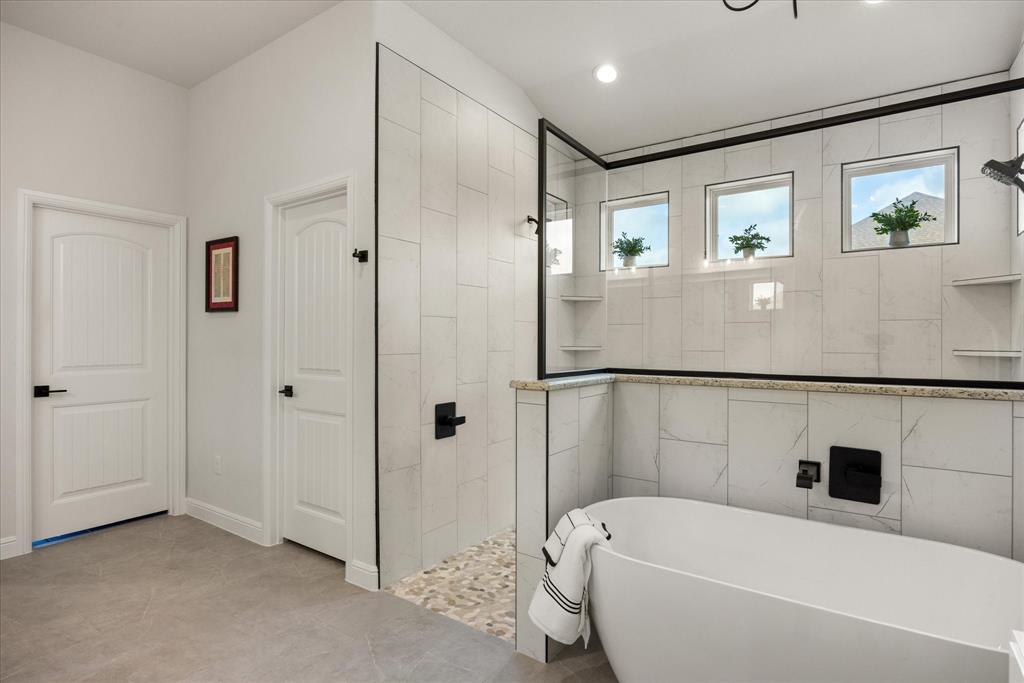 Freestanding soaking tub with matte black wall-mounted faucet, walk-in shower featuring a pebble-stone floor, glass enclosure with black framing, and neutral tile wall surrounds