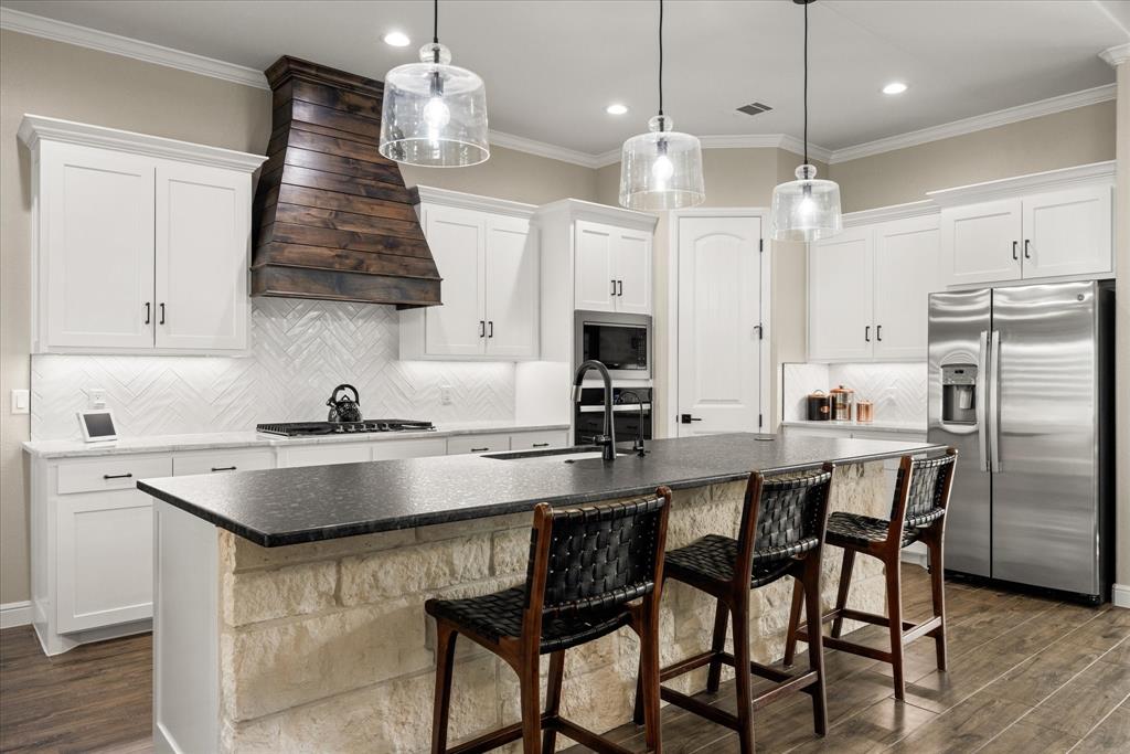 Kitchen featuring white cabinetry, a natural stone-faced island with a dark countertop, a custom dark wood range hood, a herringbone-patterned tile backsplash, and stainless steel appliances