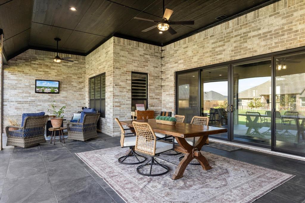 Covered outdoor patio featuring light-toned brick masonry, dark-stained wood-plank ceiling, large-format tile flooring, dual ceiling fans, and expansive sliding glass doors
