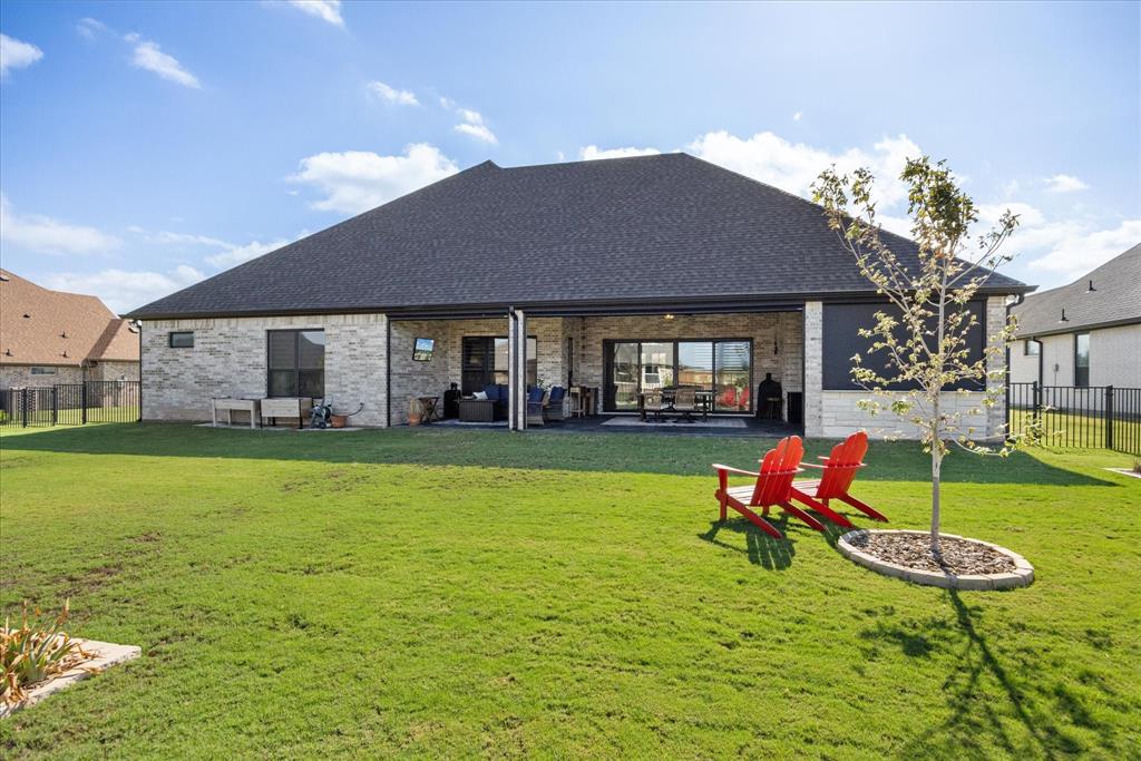 Rear exterior featuring a stone masonry facade, covered patio, dark shingle roof, and a fenced backyard with a landscaped tree island