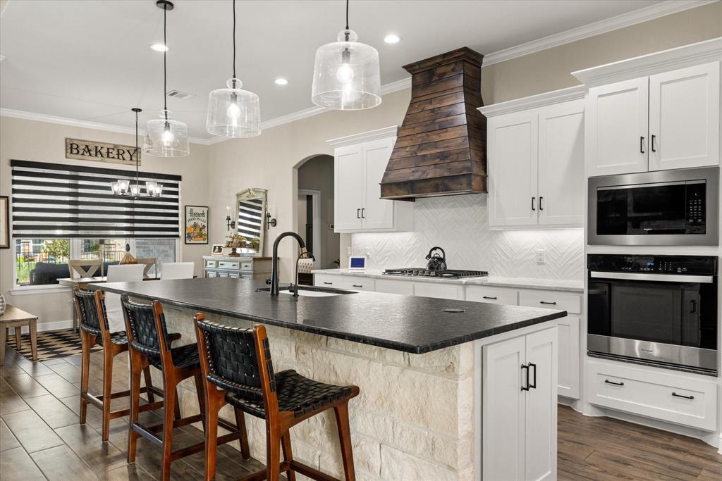 Kitchen featuring an expansive center island with a stone base and dark countertop, white cabinetry, herringbone-patterned backsplash, wood-finish vent hood, and built-in stainless steel double ovens
