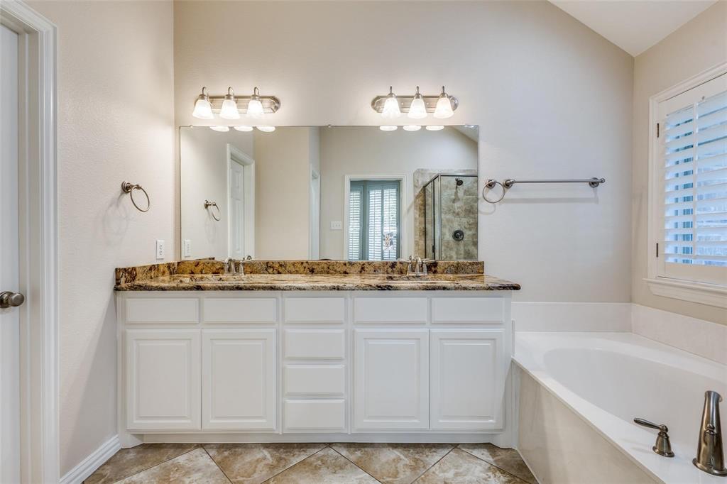 Dual vanity featuring white cabinetry, stone-surface countertop, dual under-mount sinks, wall-to-wall mirror with dual three-light fixtures, and a soaking tub with tile flooring