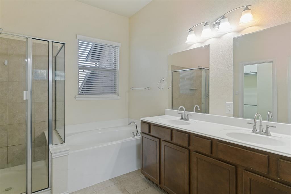 Bathroom featuring a dual-sink vanity with dark-finish cabinetry, a separate soaking tub, a glass-enclosed shower with neutral-tone wall tile, and wall-mounted vanity lighting