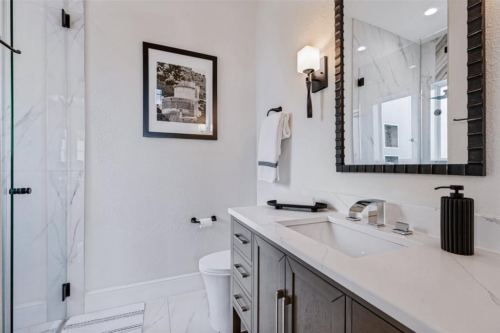 Bathroom featuring a vanity with quartz countertop, dark wood-finish cabinetry, a glass-enclosed shower with stone-patterned tile, and wall-mounted light fixtures