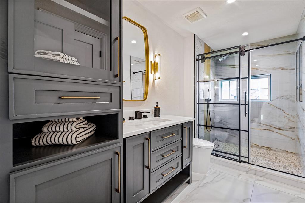 Custom gray cabinetry with brass-finish hardware, quartz countertop, arched wall-mounted mirror, and a walk-in shower featuring a sliding black-frame glass door and stone-style flooring