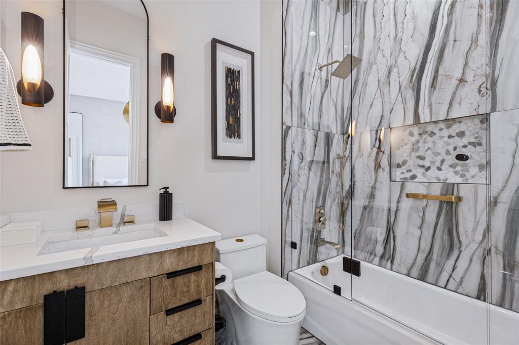 Bathroom featuring a wood-finish vanity with a rectangular sink, wall-mounted gold-tone faucet, arched mirror, and floor-to-ceiling veined tile shower surround with a built-in recessed niche