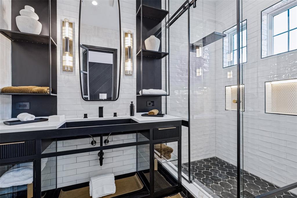 Custom metal-framed vanity with integrated shelving, quartz countertop, rectangular basin, wall-mounted vertical light fixtures, and glass-enclosed shower with tiled niches