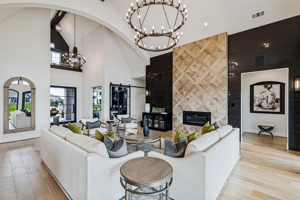 Vaulted ceiling with arched structural detailing, floor-to-ceiling fireplace featuring geometric patterned tile inlay, black tile accent wall, wide-plank wood-finish flooring, and large double-tier circular chandelier Vaulted ceiling with arched structural detailing, floor-to-ceiling fireplace featuring geometric patterned tile inlay, black tile accent wall, wide-plank wood-finish flooring, and large double-tier circular chandelier
