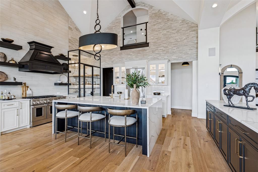Vaulted ceiling featuring a floor-to-ceiling stone accent wall, center island with stone countertop, professional-grade range with custom hood, glass-enclosed pantry, and wood-finish flooring