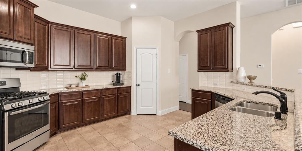 Kitchen featuring dark wood cabinetry, stone countertops, stainless steel appliances, a tile backsplash, and a double-basin undermount sink with a pull-down faucet