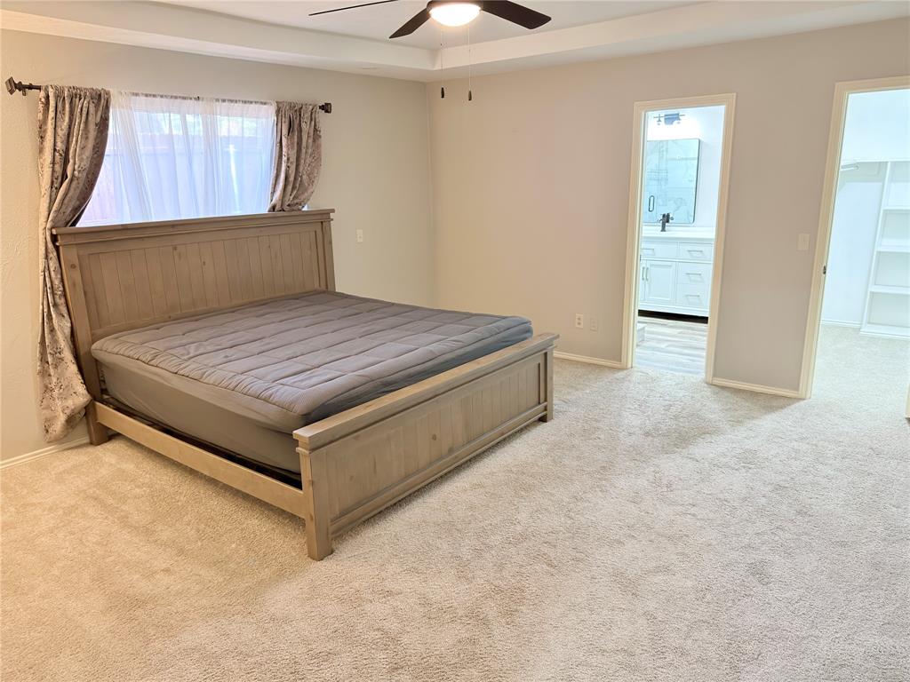 Bedroom featuring a tray ceiling, wall-to-wall carpeting, and a ceiling fan, with entryways revealing an en-suite bathroom vanity and a built-in shelving unit Bedroom featuring a tray ceiling, wall-to-wall carpeting, and a ceiling fan, with entryways revealing an en-suite bathroom vanity and a built-in shelving unit