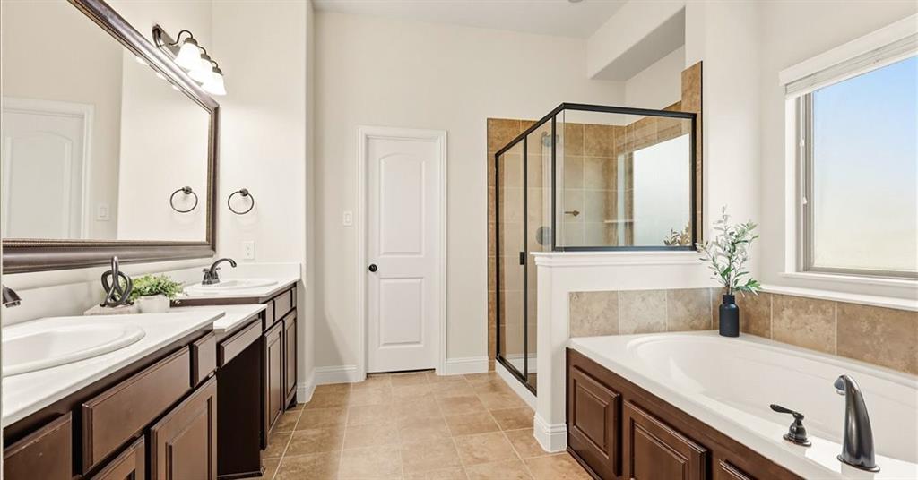Double vanity with dark wood-finish cabinetry, soaking tub with tile surround, glass-enclosed shower, large framed wall mirror, and tile flooring