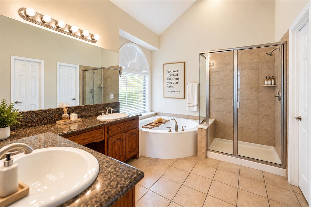 Bathroom featuring a dual-sink vanity with stone countertops, a corner soaking tub, a glass-enclosed shower, tile flooring, and an arched window  *Virtually staged to show potential*