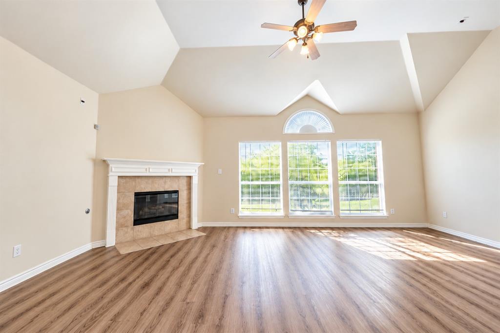 Vaulted ceiling with an integrated multi-light ceiling fan, wood-look LVP flooring, and a fireplace with a white mantel and tile surround, accented by large windows featuring a semi-circular transom