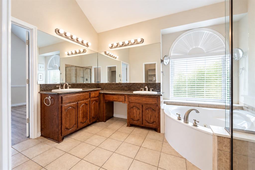 Dual-sink vanity with stone countertop, tile flooring, corner soaking tub, and arched window with integrated shutters