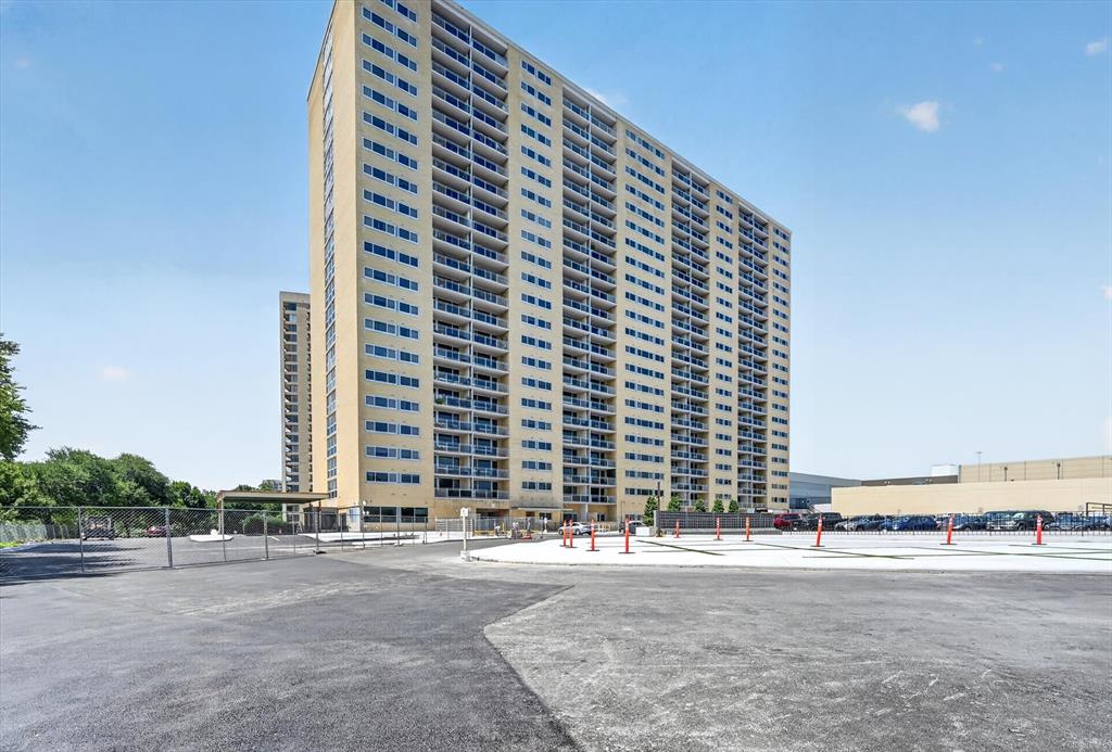 High-rise exterior featuring stacked balconies, expansive paved parking surface, and metal perimeter fencing