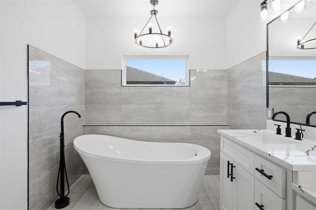Freestanding soaking tub with a floor-mounted matte black faucet, vanity with white cabinetry and a stone-surface countertop, matte black hardware, and large-format gray wall tile