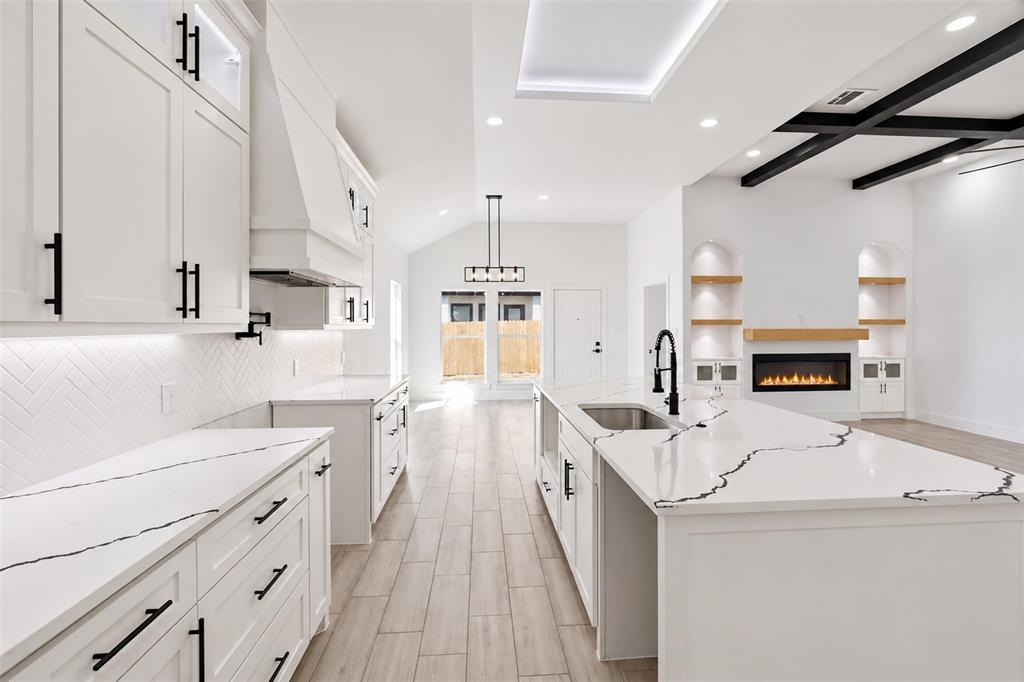 Kitchen featuring a waterfall-edge island with veined stone countertop, herringbone-pattern tile backsplash, wood-finish flooring, coffered ceiling detail, and built-in arched shelving with a fireplace