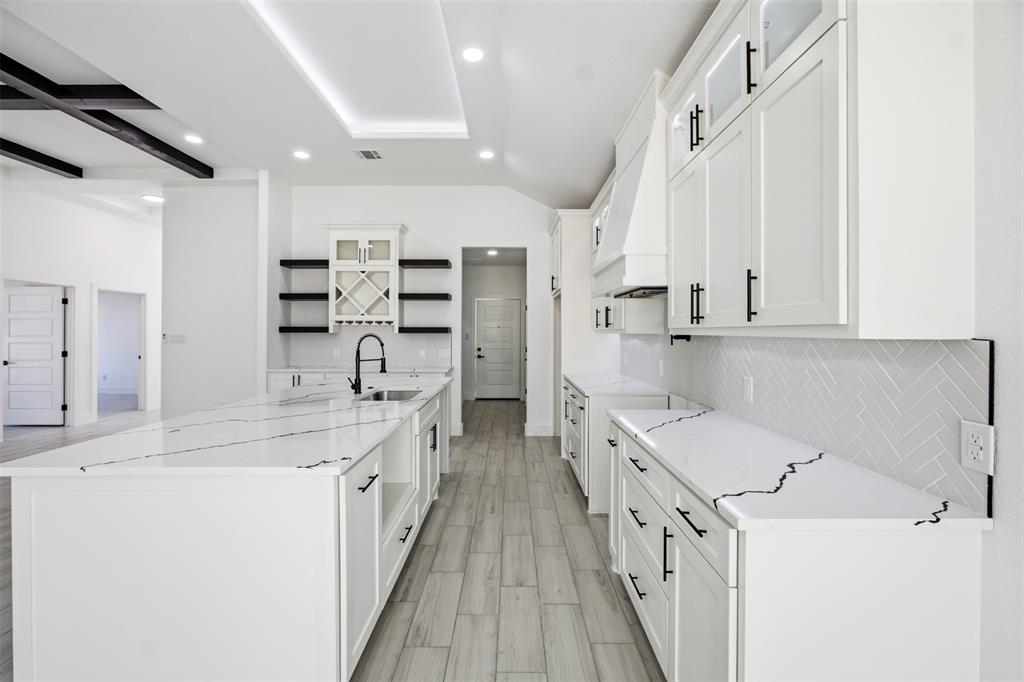 Kitchen featuring a large central island with veined stone-finish countertops, wood-finish flooring, white shaker-style cabinetry with matte black hardware, herringbone tile backsplash, and built-in wine storage
