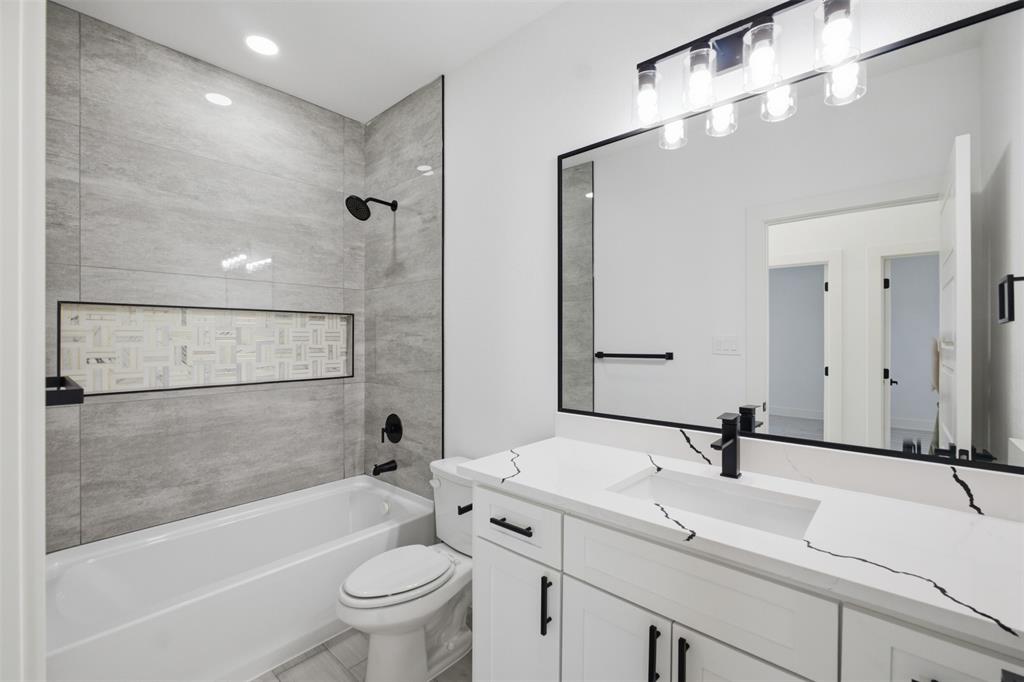 Bathroom featuring a white vanity with veined stone-finish countertop, undermount sink, matte black hardware, tiled tub surround with recessed decorative niche, and a large framed mirror with multi-bulb vanity lighting