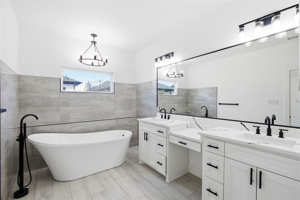Freestanding soaking tub with matte black floor-mounted faucet, dual-sink vanity with white stone-finish countertop, full-width vanity mirror, tile-surround wall detailing, and wood-finish flooring