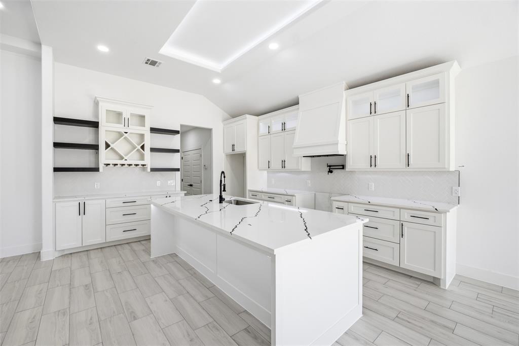 Kitchen featuring white shaker cabinetry, a waterfall-edge center island with stone-patterned surfacing, integrated wine storage, a farmhouse-style range hood, and wood-finish flooring