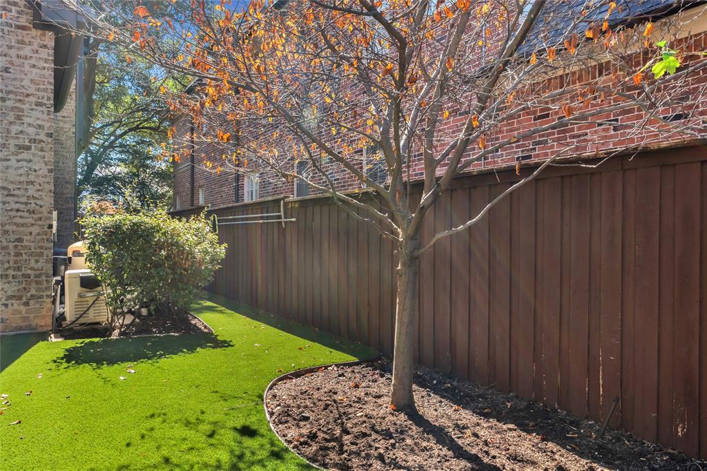 Exterior space featuring a wood-panel privacy fence, synthetic turf lawn, and a mature tree within a landscaped garden bed