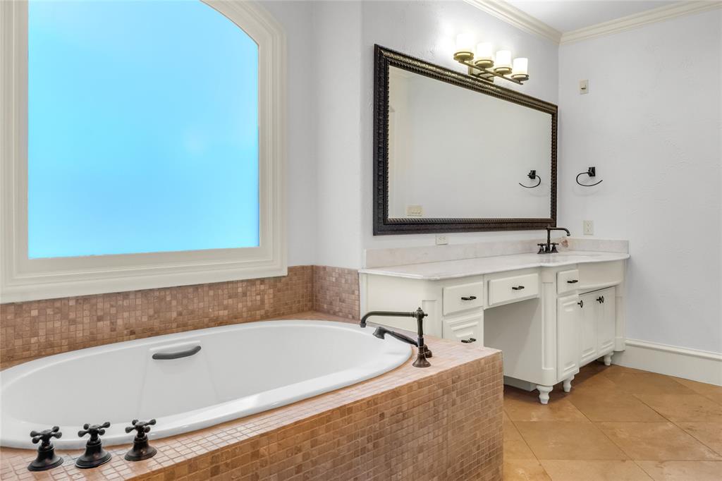 Soaking tub with mosaic tile surround, built-in vanity with stone countertop, oversized framed mirror, and arched window