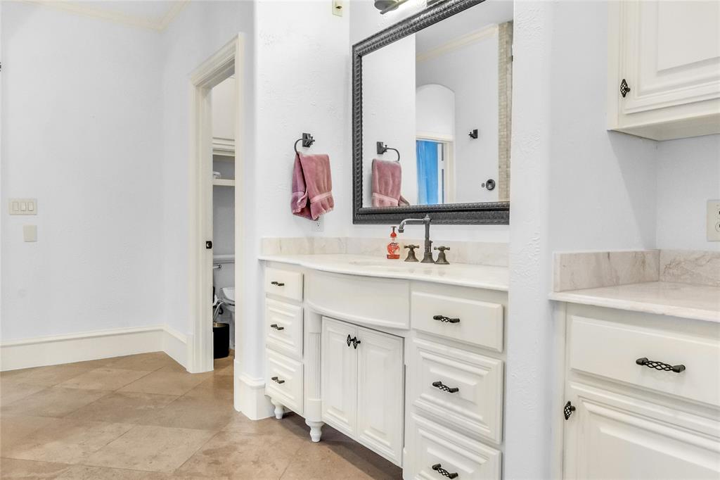 Custom vanity featuring white cabinetry, stone countertop with integrated sink, oil-rubbed bronze hardware, framed wall-mounted mirror, and large-format tile flooring