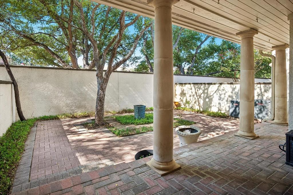 Covered brick patio featuring structural stone columns, perimeter stucco wall, herringbone brick pavers, and mature trees