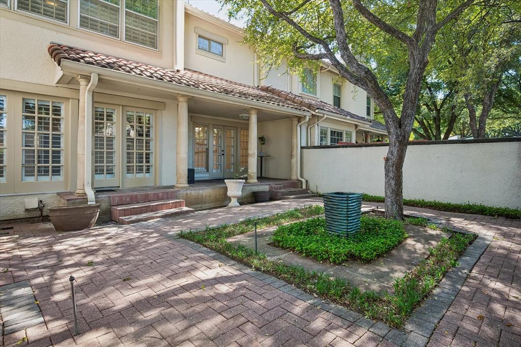 Exterior patio ad courtyard featuring patterned brick pavers, covered porch with classical columns, stucco siding, and a mature tree within a landscaped garden bed Exterior patio ad courtyard featuring patterned brick pavers, covered porch with classical columns, stucco siding, and a mature tree within a landscaped garden bed