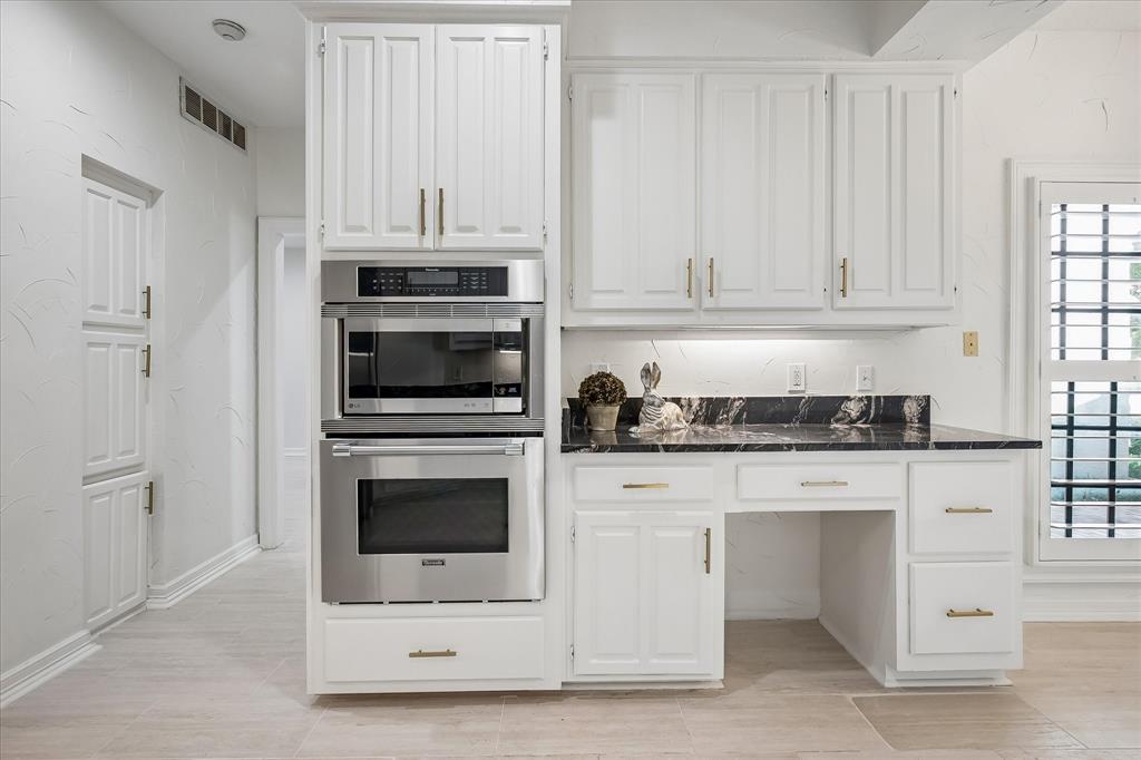 Kitchen cabinetry featuring white shaker-style doors, brass-finish hardware, dark stone-finish countertops, stainless steel built-in double ovens, and a dedicated workspace area