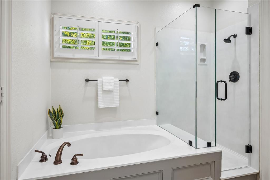 Oversized soaking tub with solid-surface surround, walk-in shower with frameless glass enclosure, matte black plumbing fixtures, and white plantation shutters