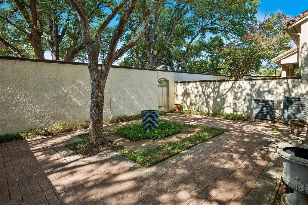 Enclosed courtyard featuring decorative brick pavers, mature trees, a central ground-level planter, and a perimeter stucco wall with a metal gate
