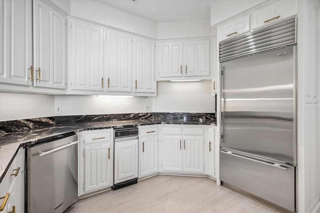 Kitchen featuring white raised-panel cabinetry with brass-finish hardware, dark stone-patterned countertops, stainless steel integrated refrigerator, built-in dishwasher, and wood-finish flooring