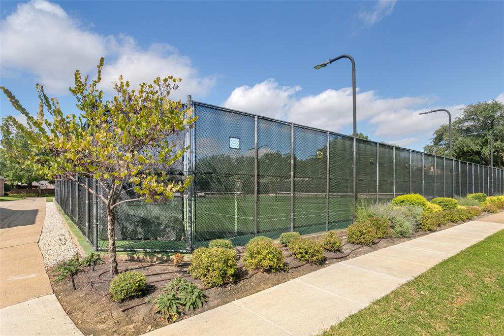 Private tennis court enclosed by chain-link fencing, featuring integrated pole-mounted lighting, concrete walkways, and landscaped shrubbery