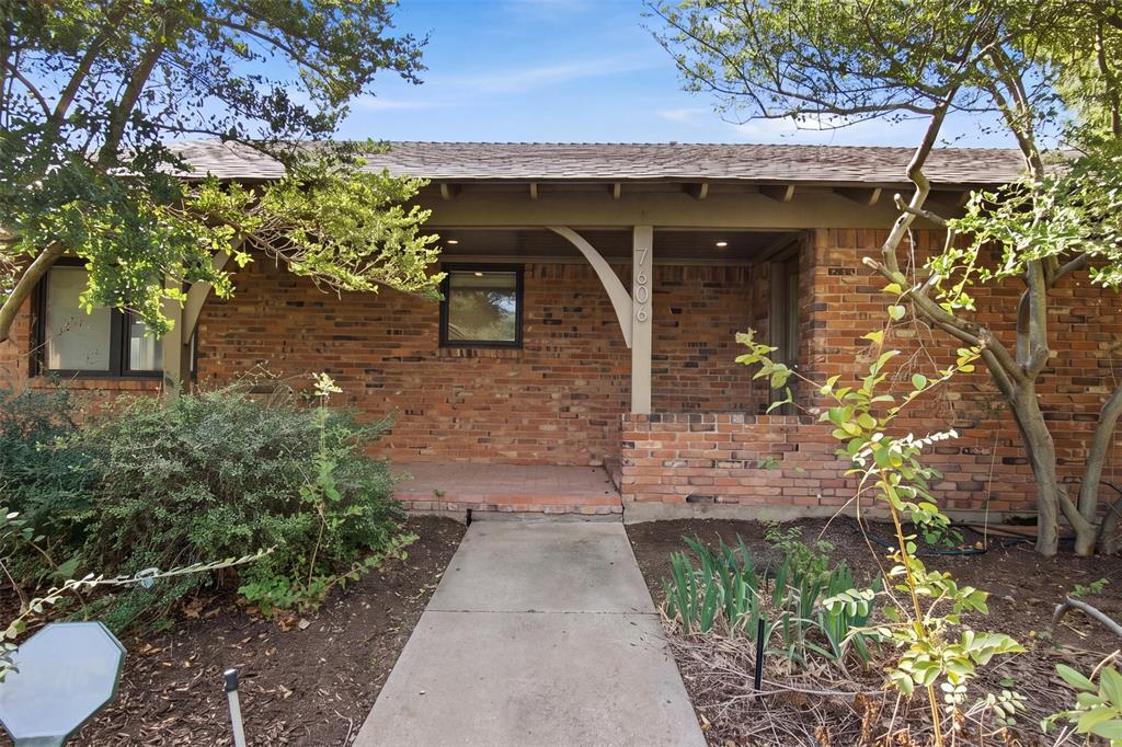 Single-story exterior featuring a brick facade, covered porch, arched support brackets, and concrete walkway