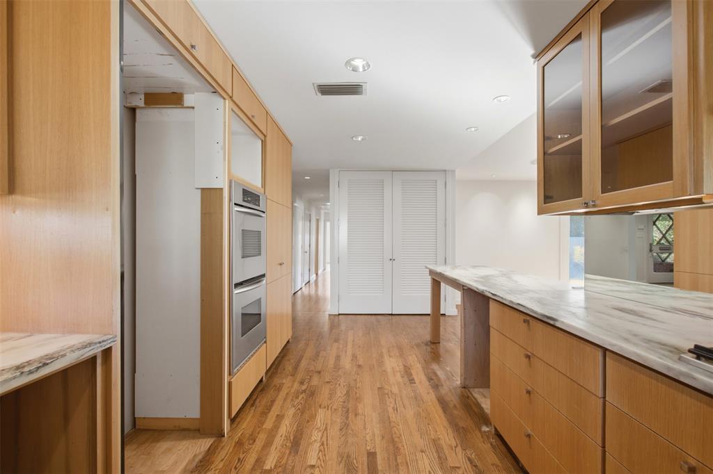 Kitchen featuring light wood-finish cabinetry, stone countertops with integrated backsplash, wood-finish flooring, built-in double wall ovens, and glass-front upper cabinets with recessed lighting