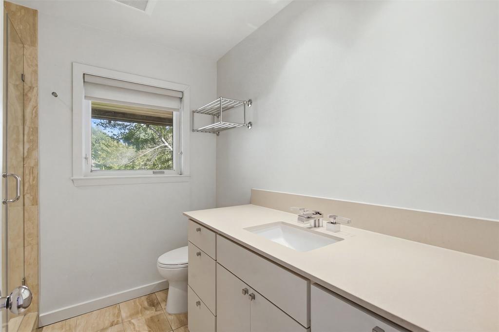 Bathroom featuring a light-toned vanity with an integrated countertop and sink, stone-tile flooring, a glass-enclosed shower with stone surround, and a wall-mounted metal shelving unit