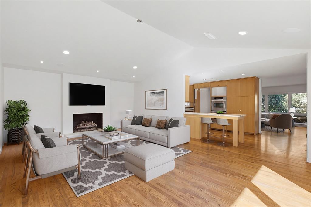 Open-concept living space featuring wood-finish flooring, vaulted ceilings, recessed lighting, a fireplace with a minimalist hearth, and light-toned cabinetry with built-in appliances.