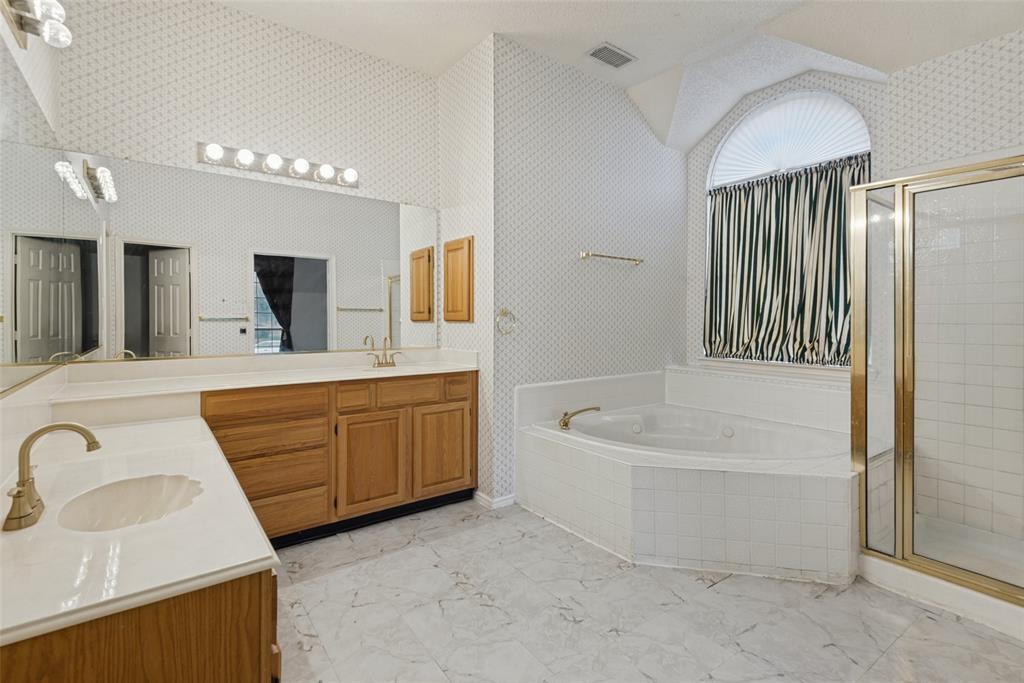 Bathroom featuring a dual-sink vanity with light-toned countertops, a large jetted corner soaking tub with a tile surround, a glass-enclosed shower, and an arched window