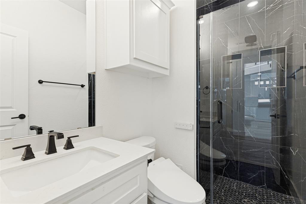 White vanity with a solid-surface countertop and integrated sink, matte black widespread faucet, walk-in shower with dark tile surround and pebble flooring, and built-in upper wall cabinetry