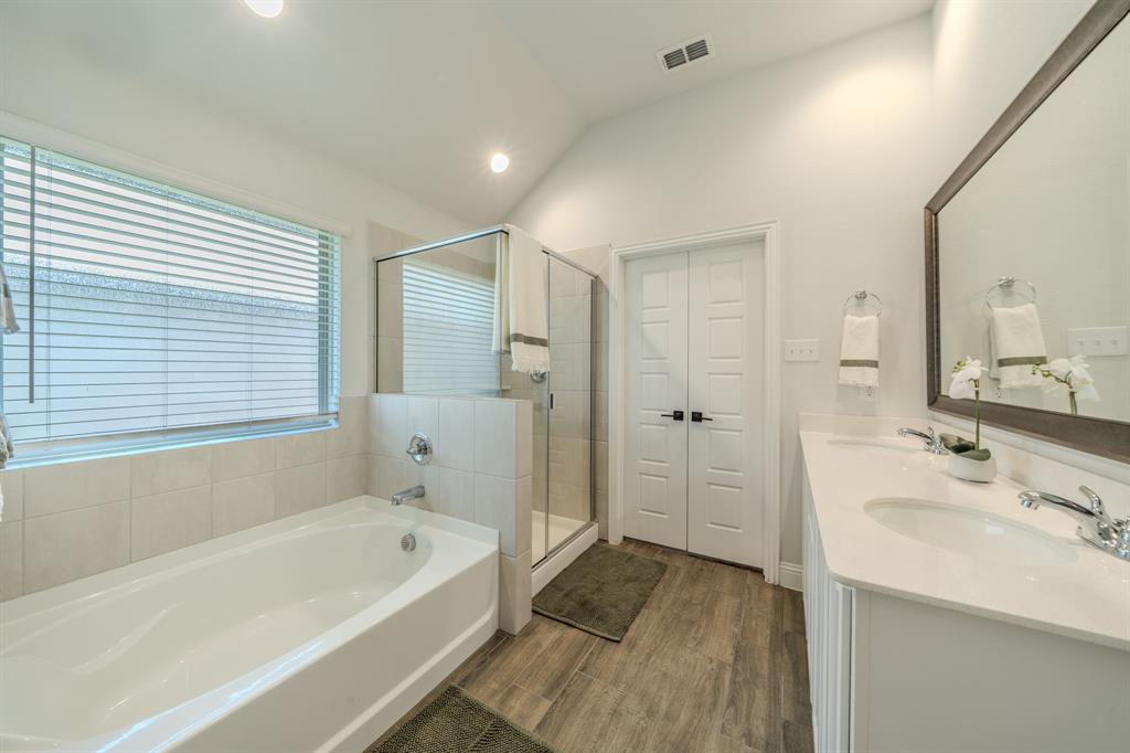 Bathroom featuring a vaulted ceiling, dual-sink vanity with light-toned countertop, glass-enclosed shower, built-in soaking tub, and wood-finish flooring