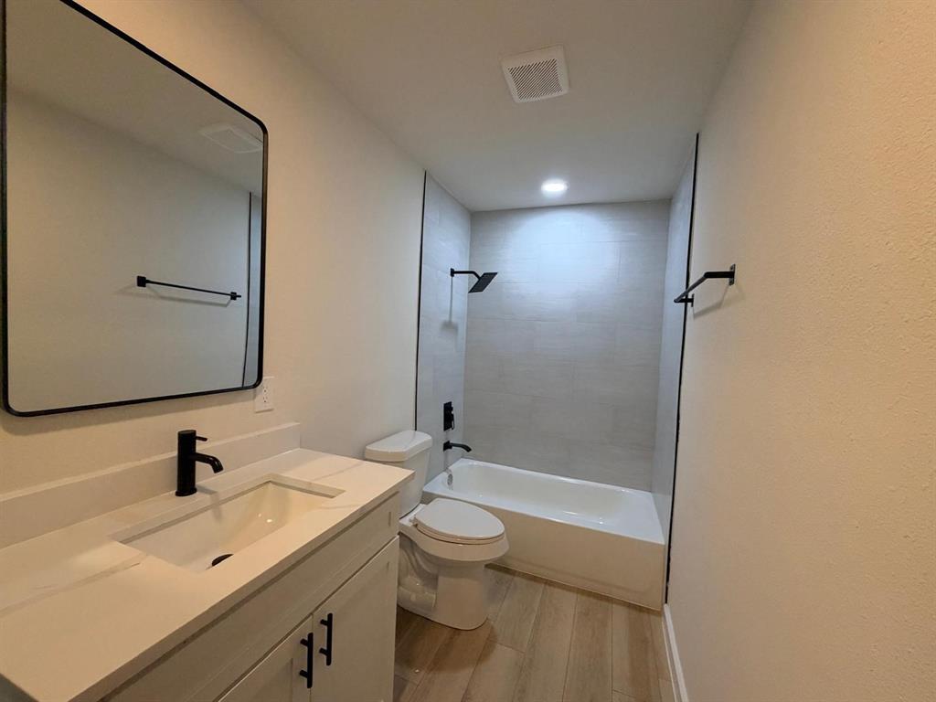 Bathroom featuring a vanity with a white countertop and rectangular undermount sink, black hardware, a framed mirror, wood-finish flooring, and a shower-tub combination with light-toned wall tile