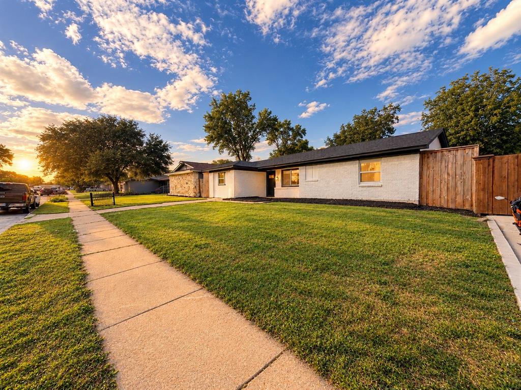 Single-story residence featuring white painted brick and stone exterior, a dark shingled roof, a front lawn, and wood privacy fencing