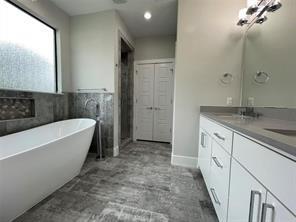 Freestanding soaking tub with floor-mounted faucet, vanity with dual undermount sinks, stone-finish tile flooring, recessed wall niche, and multi-light vanity fixture