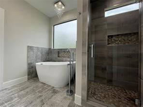 Freestanding soaking tub with a floor-mounted faucet, walk-in shower with stone tile flooring and recessed shelving, wood-finish tile flooring, and a crystal chandelier