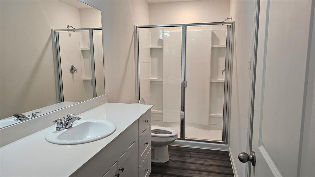 Bathroom featuring a white vanity with an integrated sink, a full-width mirror, wood-finish flooring, and a walk-in shower with a glass enclosure and built-in shelving