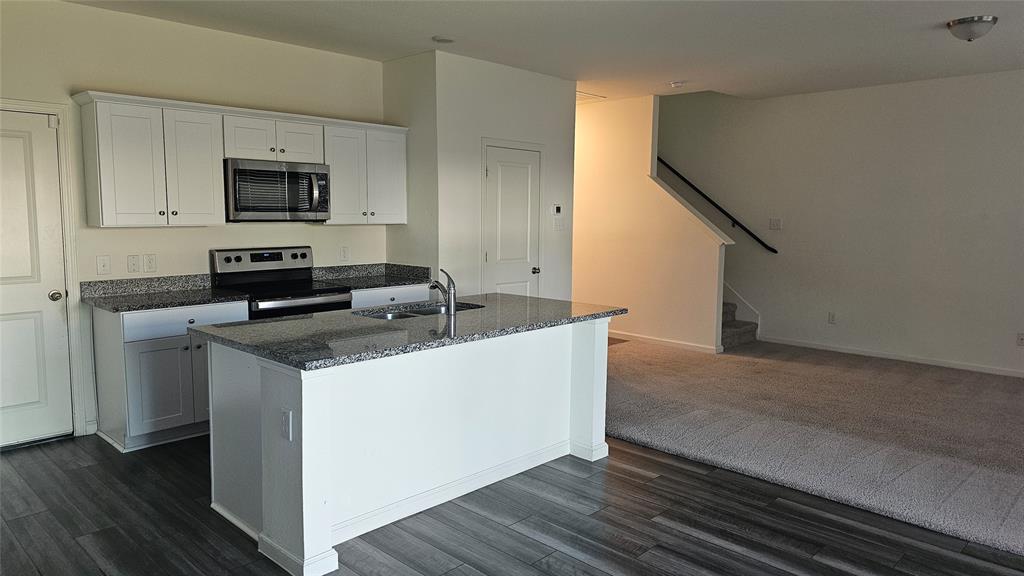 Kitchen featuring white cabinetry, stone-finish countertops, stainless steel appliances, a center island with a dual-basin sink, and wood-finish flooring Kitchen featuring white cabinetry, stone-finish countertops, stainless steel appliances, a center island with a dual-basin sink, and wood-finish flooring