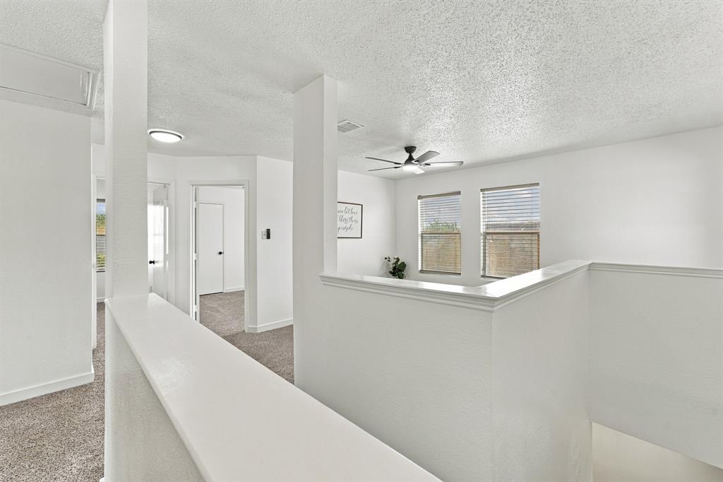 Open floor plan interior featuring textured ceilings, carpeted flooring, a multi-blade ceiling fan with integrated lighting, and dual windows with horizontal blinds